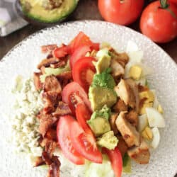 a cobb salad on a white plate next to two tomatoes