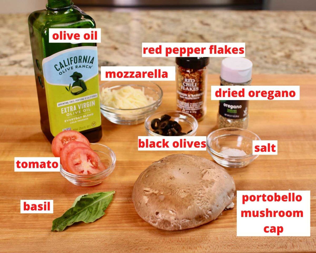 olive oil, cheese, spices, olives, tomato slices, a basil leaf and one portobello mushroom cap on a brown cutting board.
