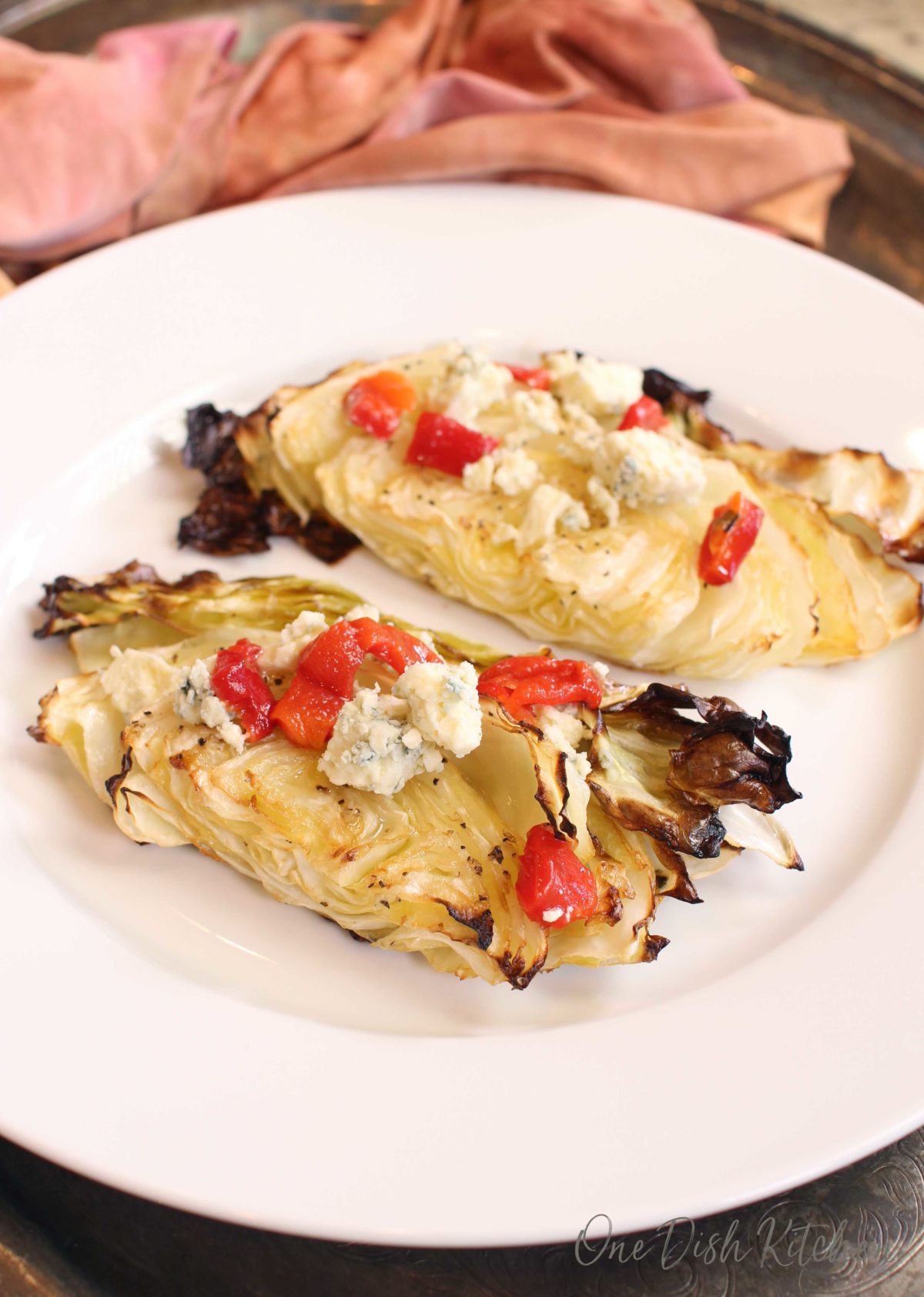 roasted cabbage topped with blue cheese and red peppers on a white plate next to an orange napkin.