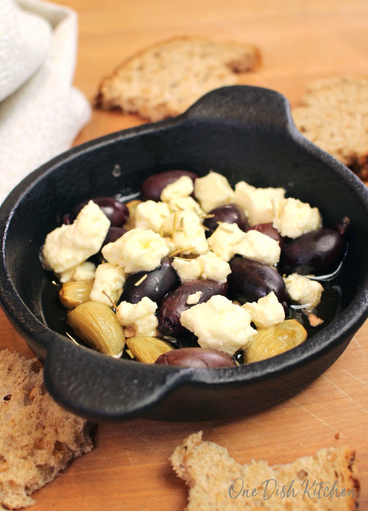 a bowl of baked feta with kalamata olives, olive oil, and garlic on a brown cutting board next to torn pieces of brown bread.