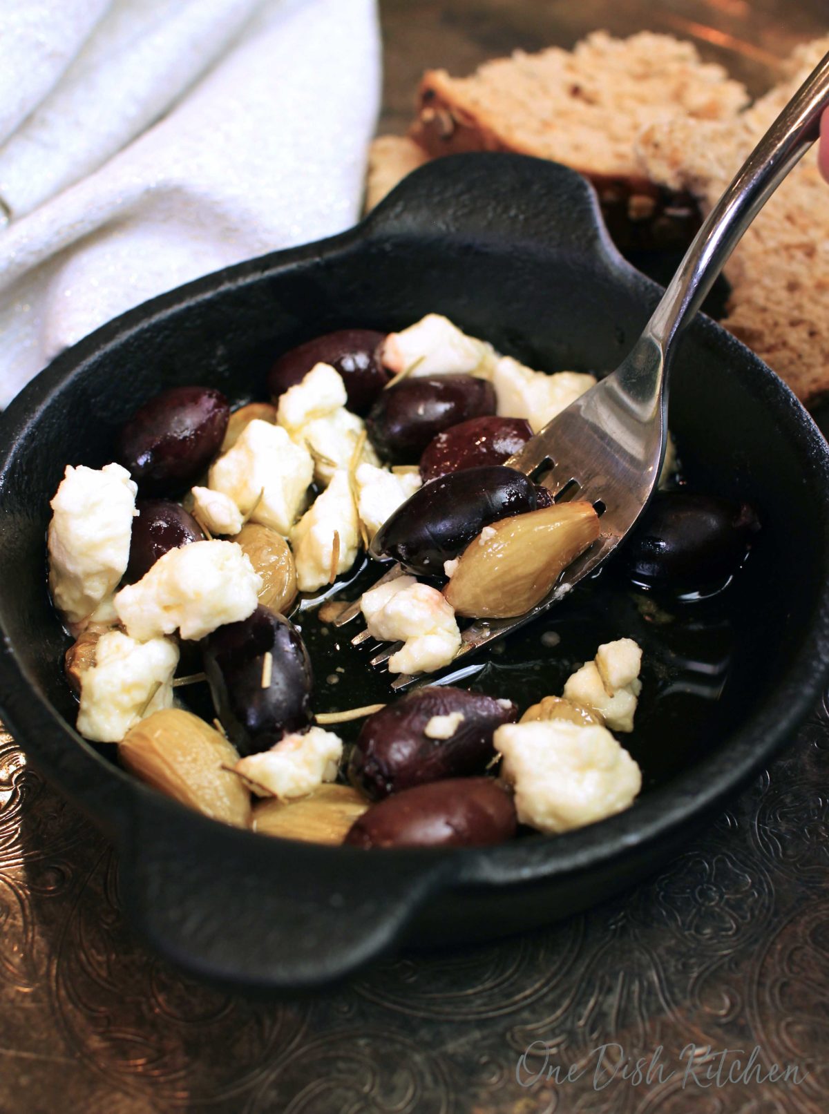 a small cast iron skillet filled with cloves of roasted garlic, olives, and feta with a fork on the side of the dish.