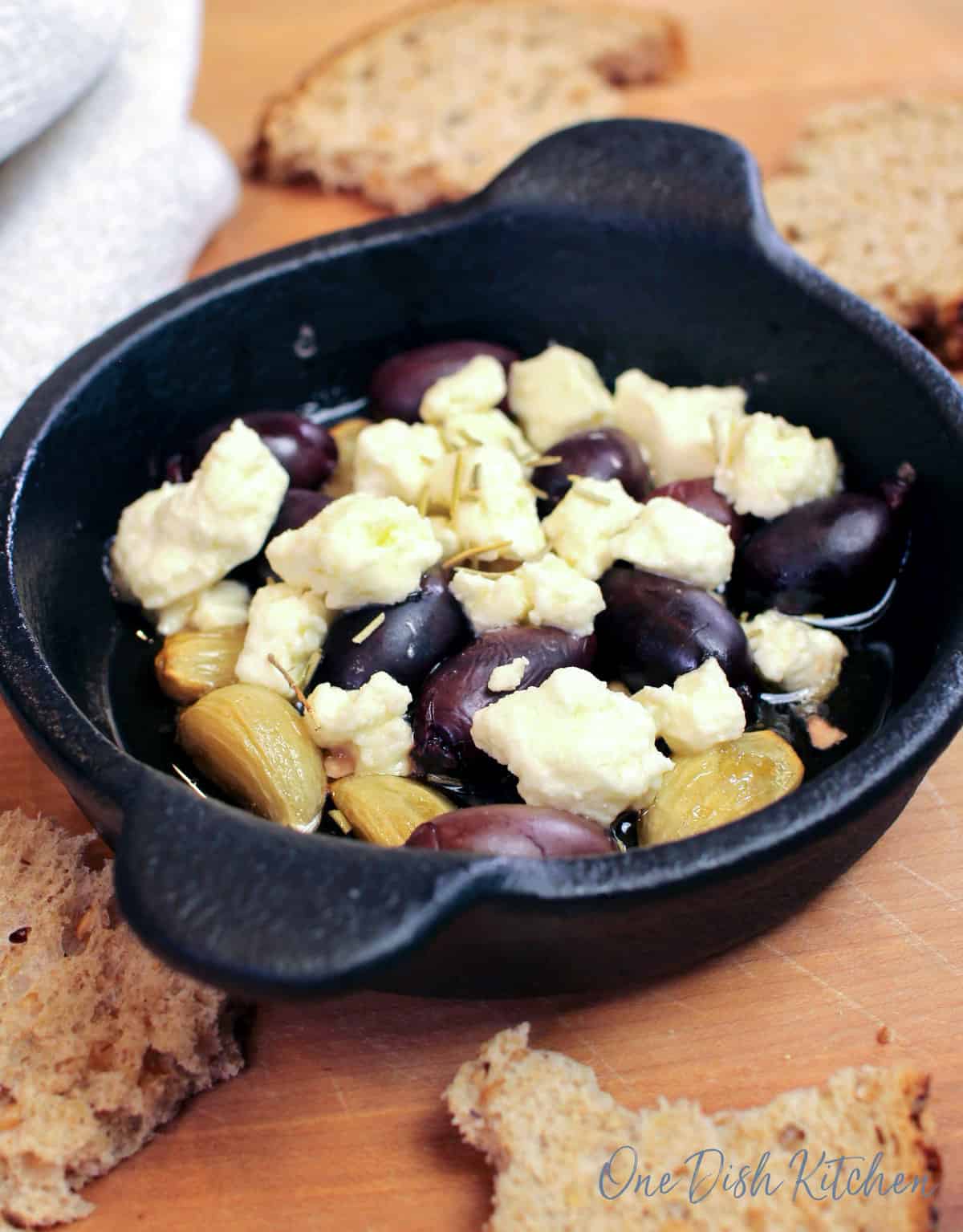 a small baking dish filled with roasted garlic, olives, and feta cheese next to torn pieces of bread.