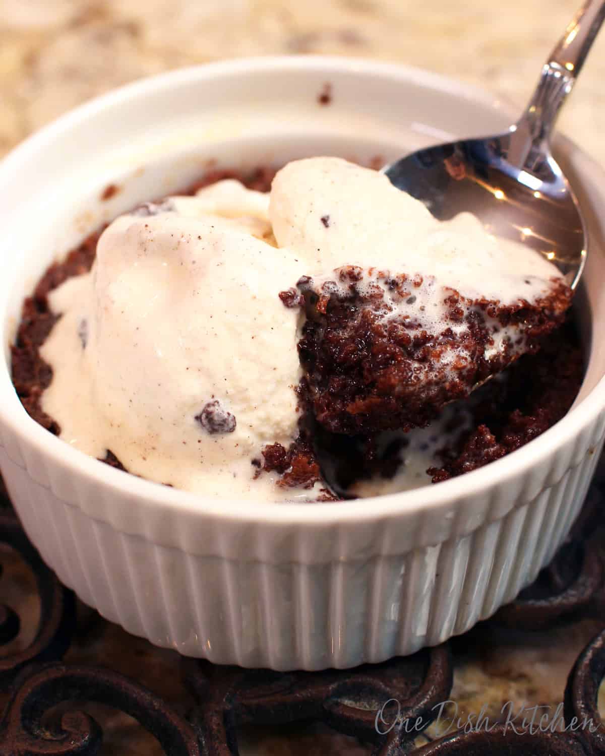 a single serve brownie baked in a ramekin topped with vanilla ice cream and a spoon digging into the dish.