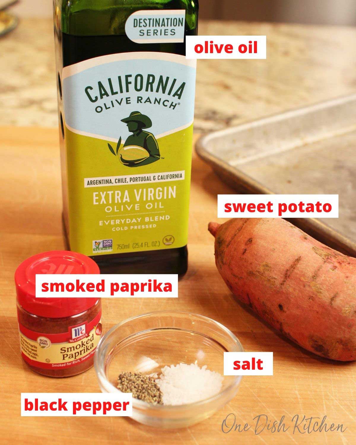 olive oil, a sweet potato, spice jar, salt and pepper on a brown cutting board.