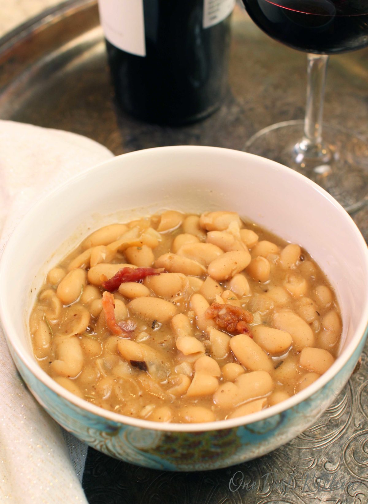 a white bowl filled with soup made with beans and bacon next to a glass of red wine and a wine bottle on a silver tray.