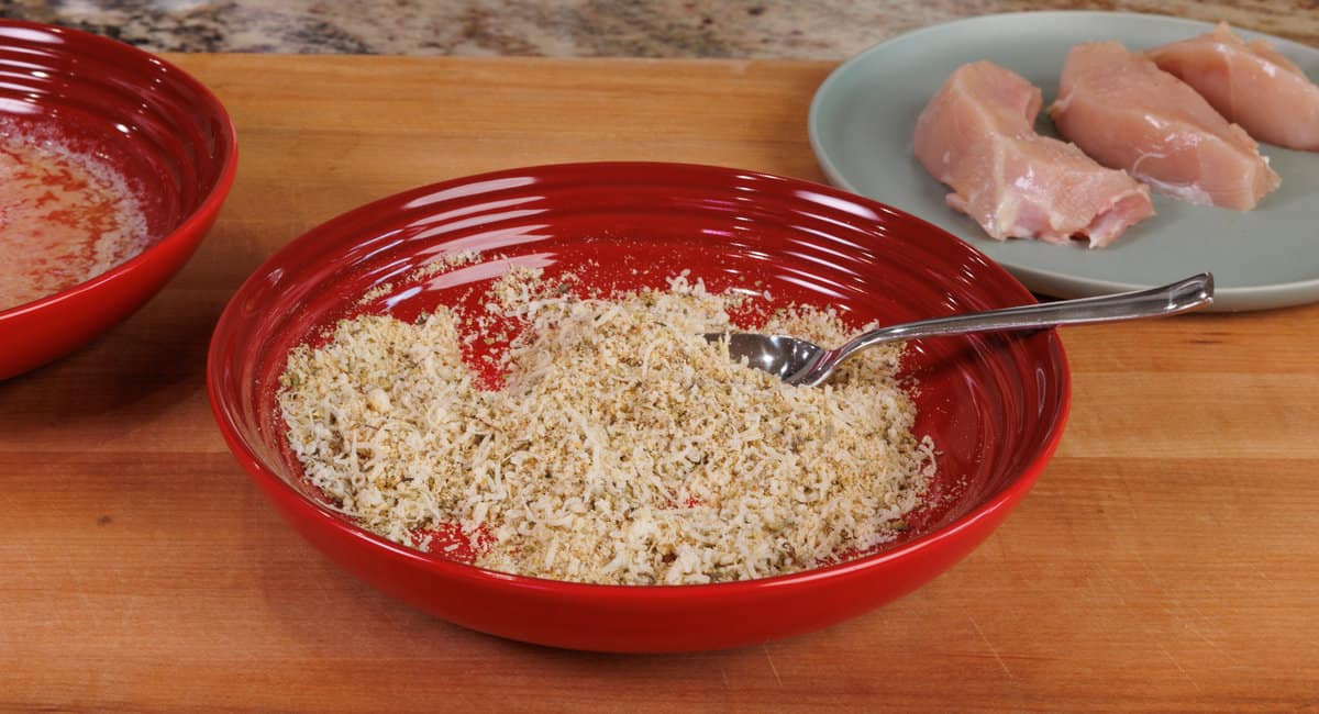 a bowl of breadcrumbs, shredded cheese,  and seasonings on a wooden cutting board