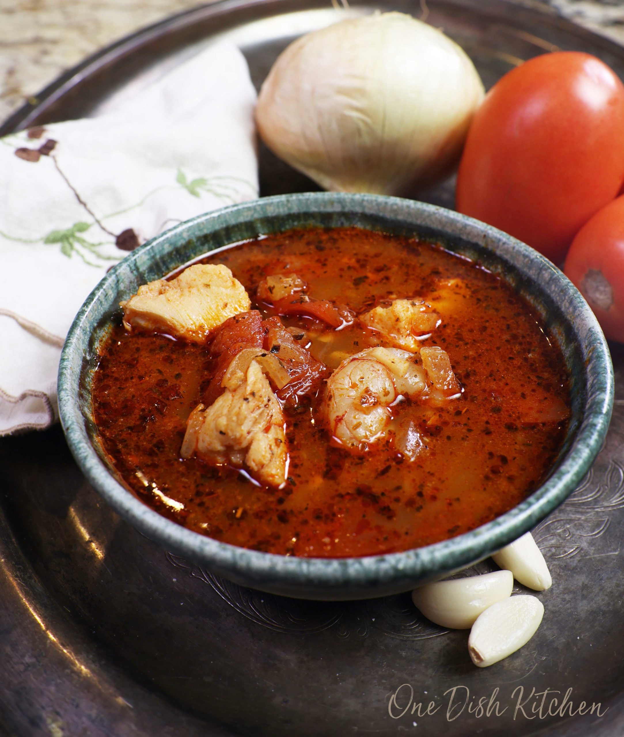 a blue bowl filled with fish stew on a silver tray next to tomatoes, garlic cloves, and an onion.