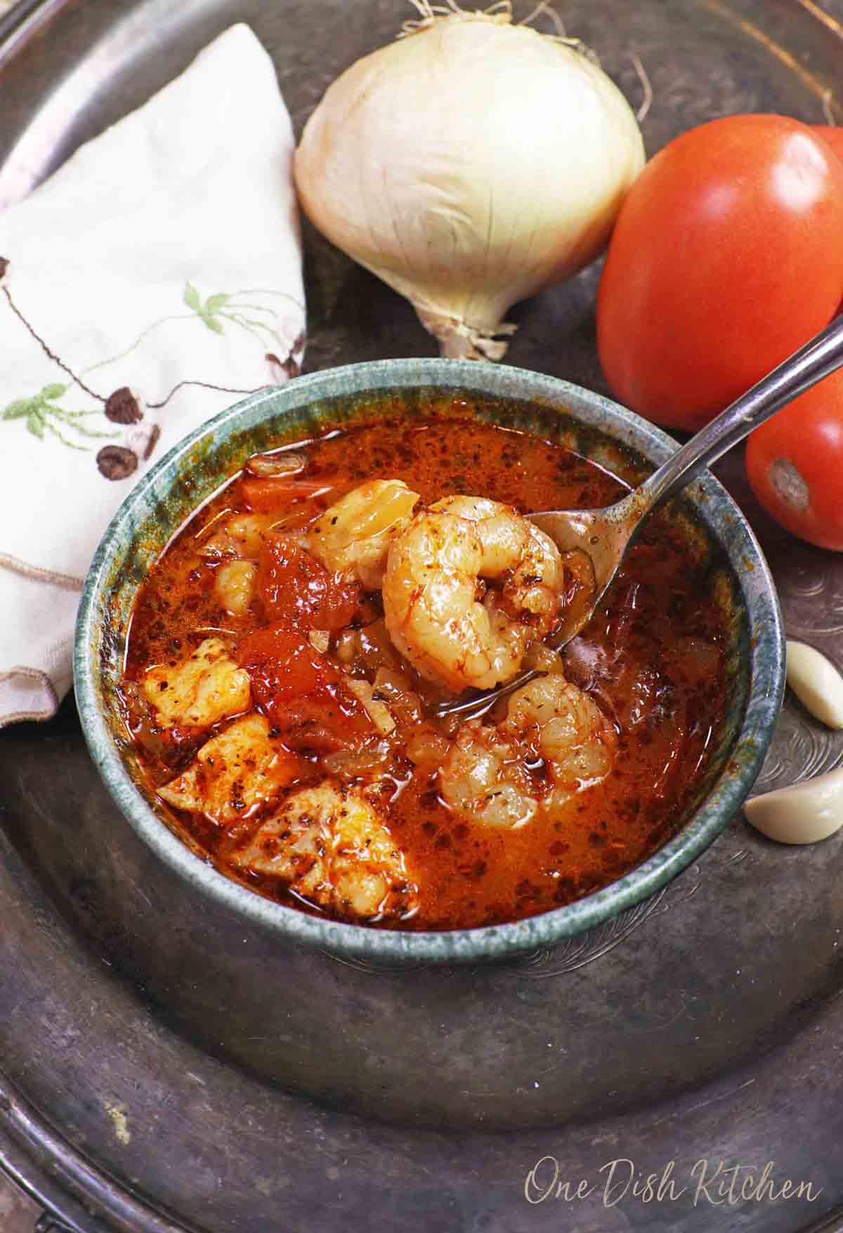 a blue bowl filled with cioppino with a large shrimp on a spoon above the dish.