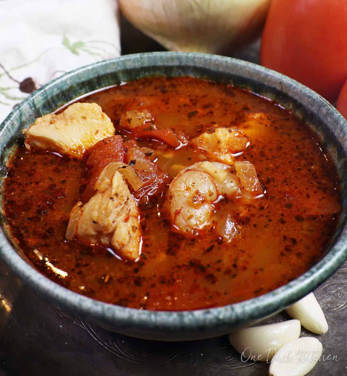 a blue bowl filled with fish stew on a silver tray next to tomatoes, garlic cloves, and an onion.