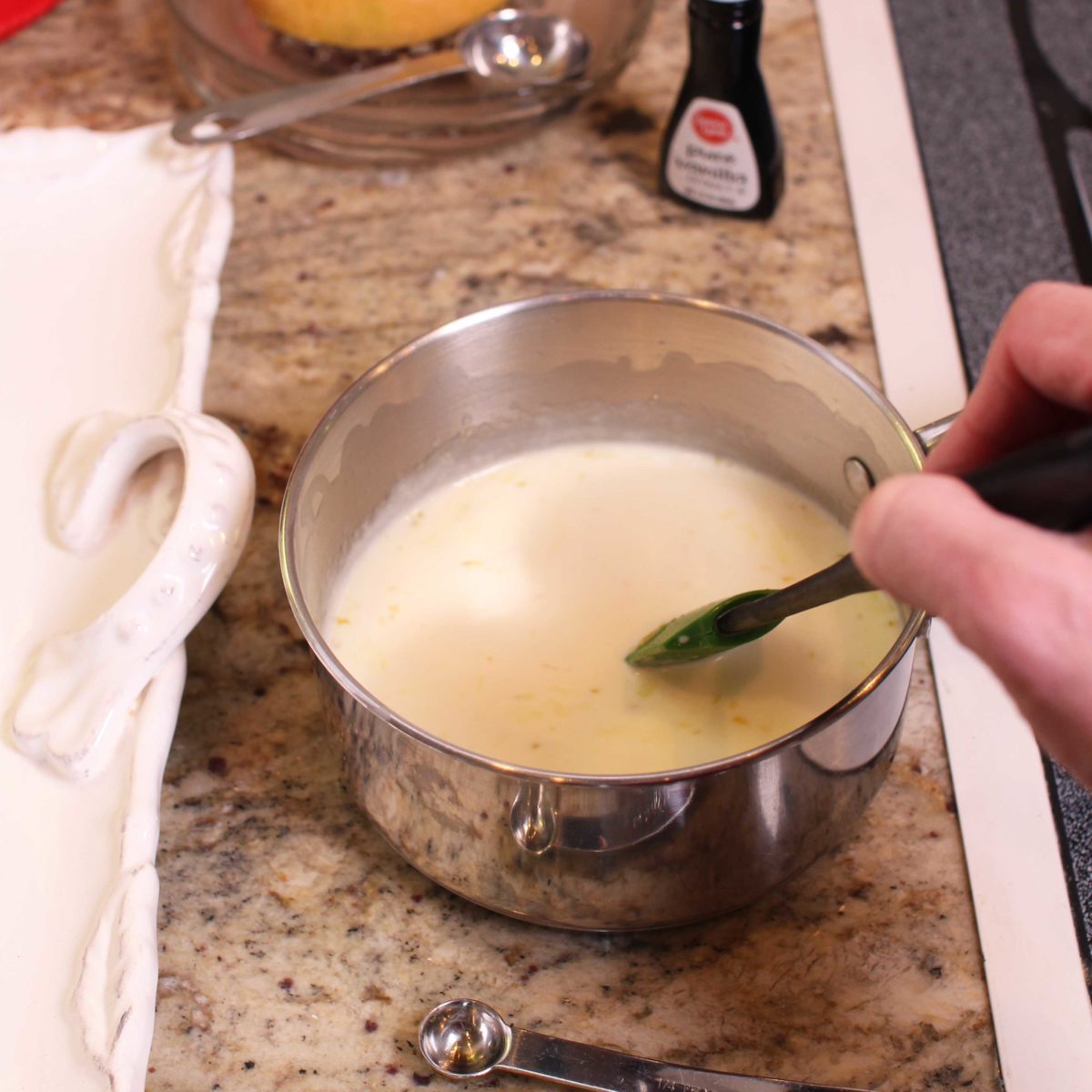 stirring a custard in a small silver saucepan next to a jar of vanilla extract.