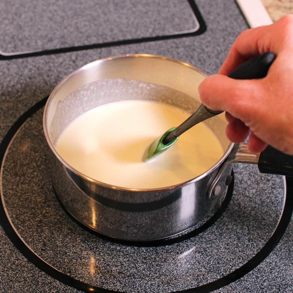 cream being stirred with a green spoon in a small silver pot