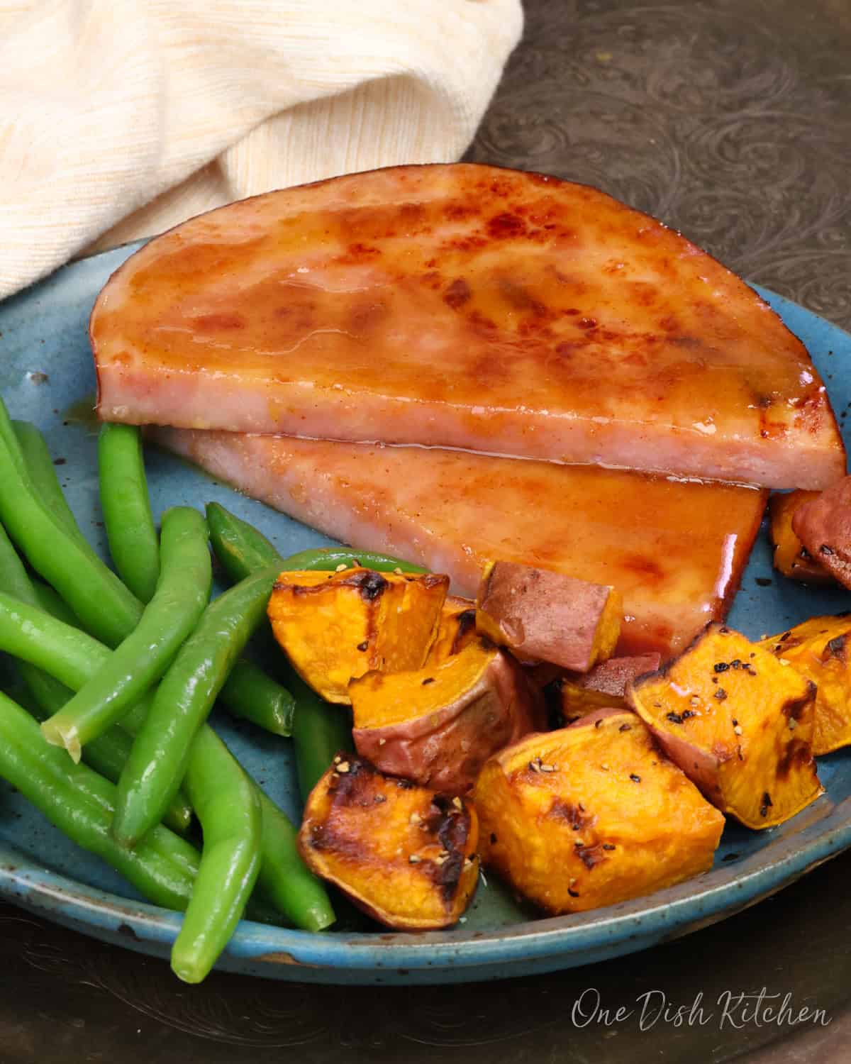 a glazed ham steak on a blue plate with roasted sweet potatoes and green beans.