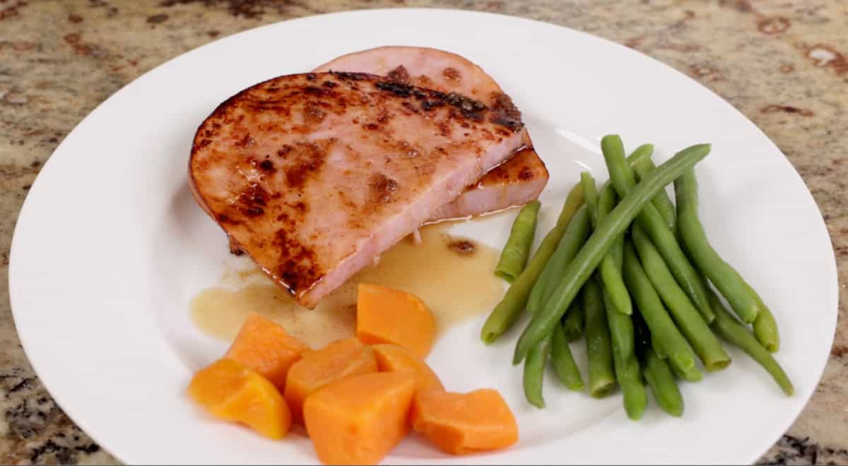 a ham steak on a white plate next to sweet potatoes and green beans.