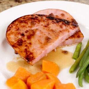 a ham steak on a white plate next to sweet potatoes and green beans.