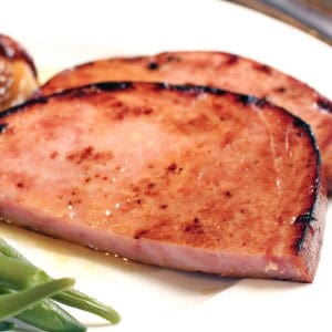 a ham steak on a white plate next to sweet potatoes and green beans.