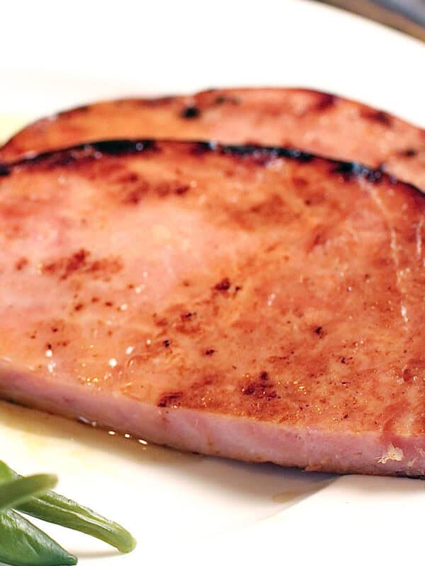 a ham steak on a white plate next to sweet potatoes and green beans.