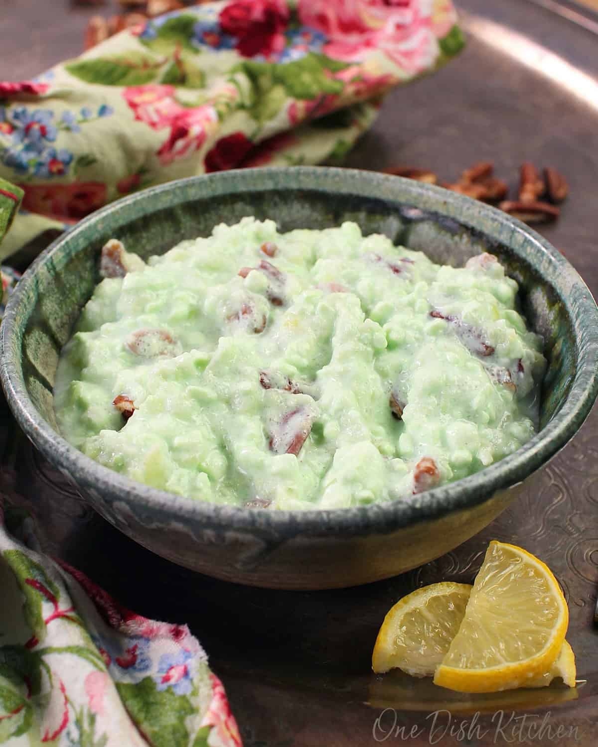 a blue bowl filled with green jello fluff topped with nuts on a silver tray next to two lemon slices.