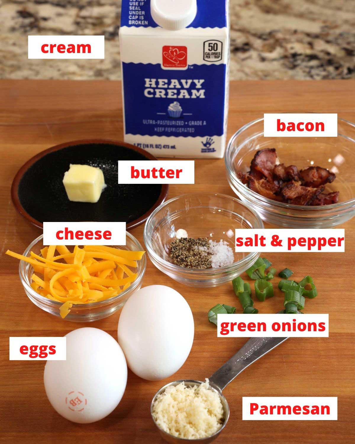 eggs, cheese, bacon, cream, butter, and green onions on a brown wooden cutting board.