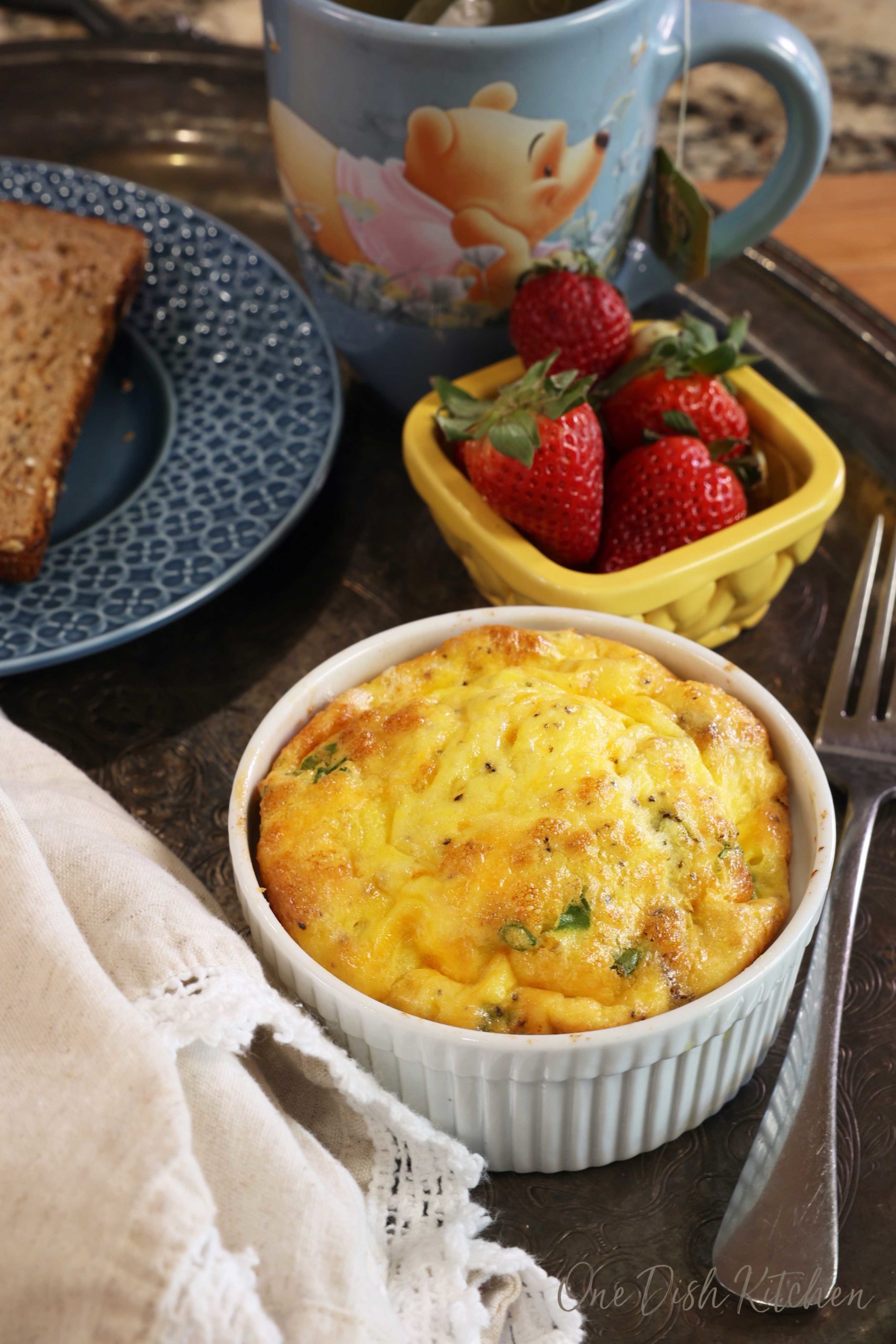 a ramekin filled with a baked egg casserole next to a bowl of strawberries and a cup of tea.