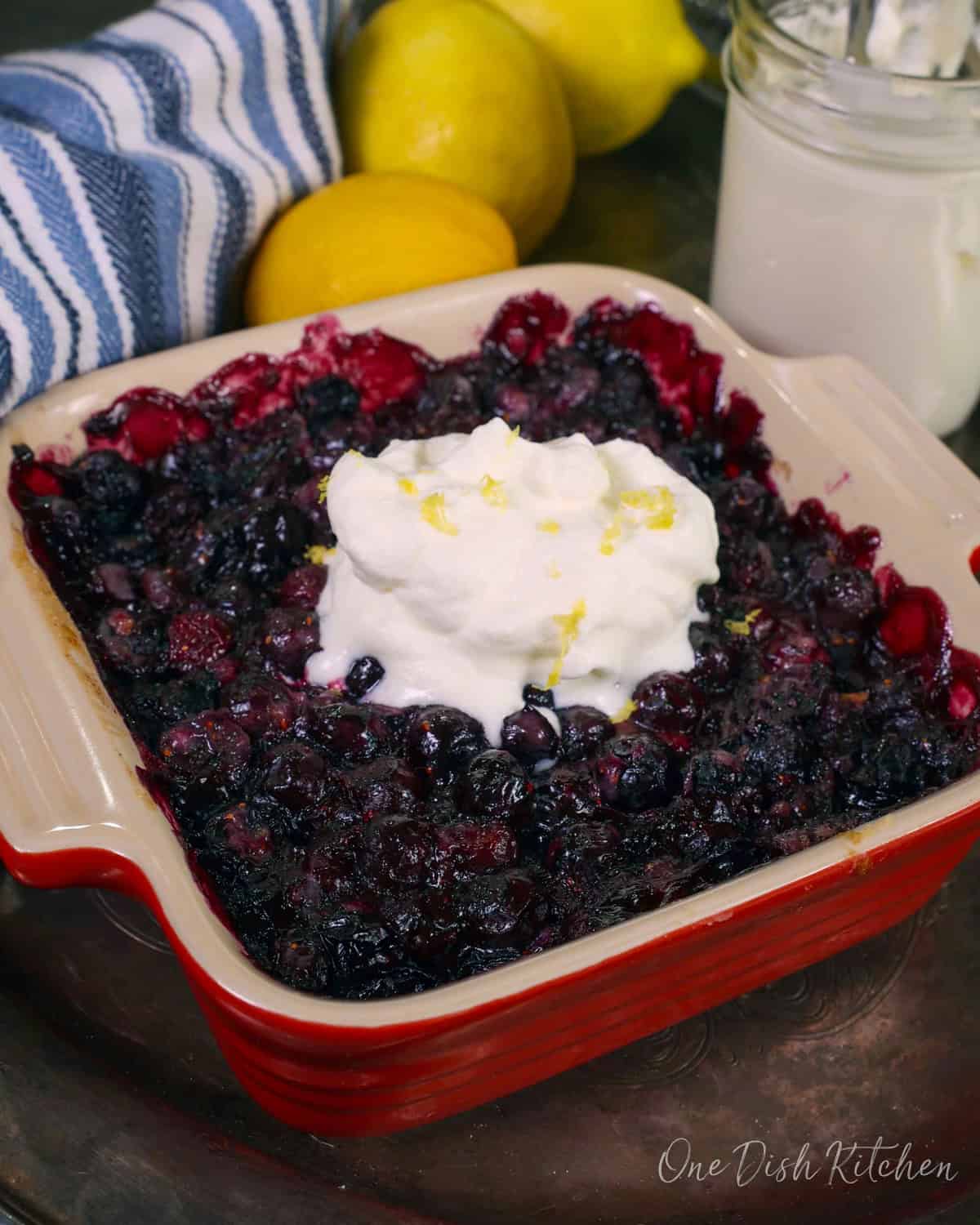 a blueberry pie baked in a square baking dish topped with whipped cream next to three lemons and a blue napkin.