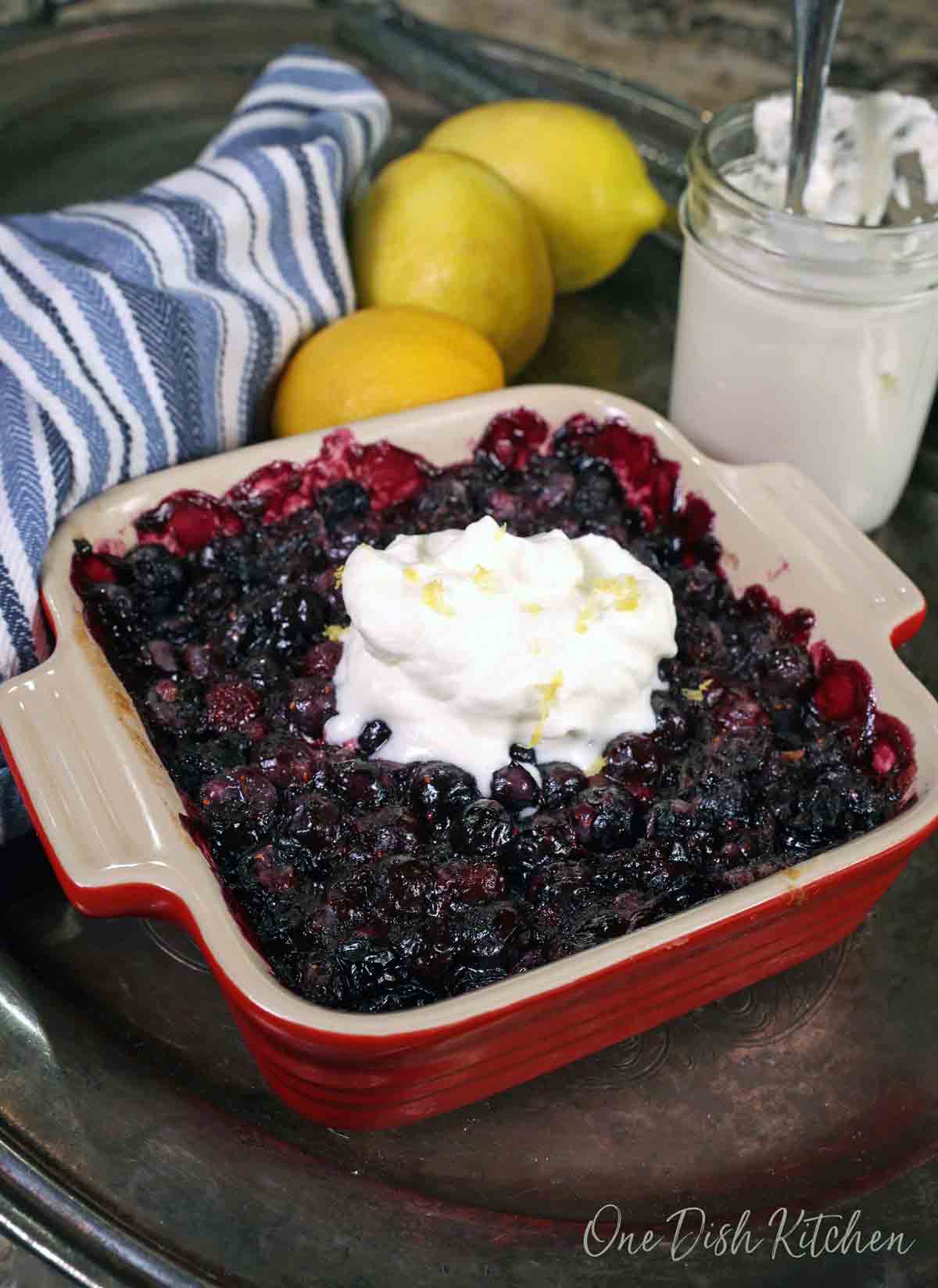 a blueberry pie baked in a square baking dish topped with whipped cream next to three lemons and a blue napkin.