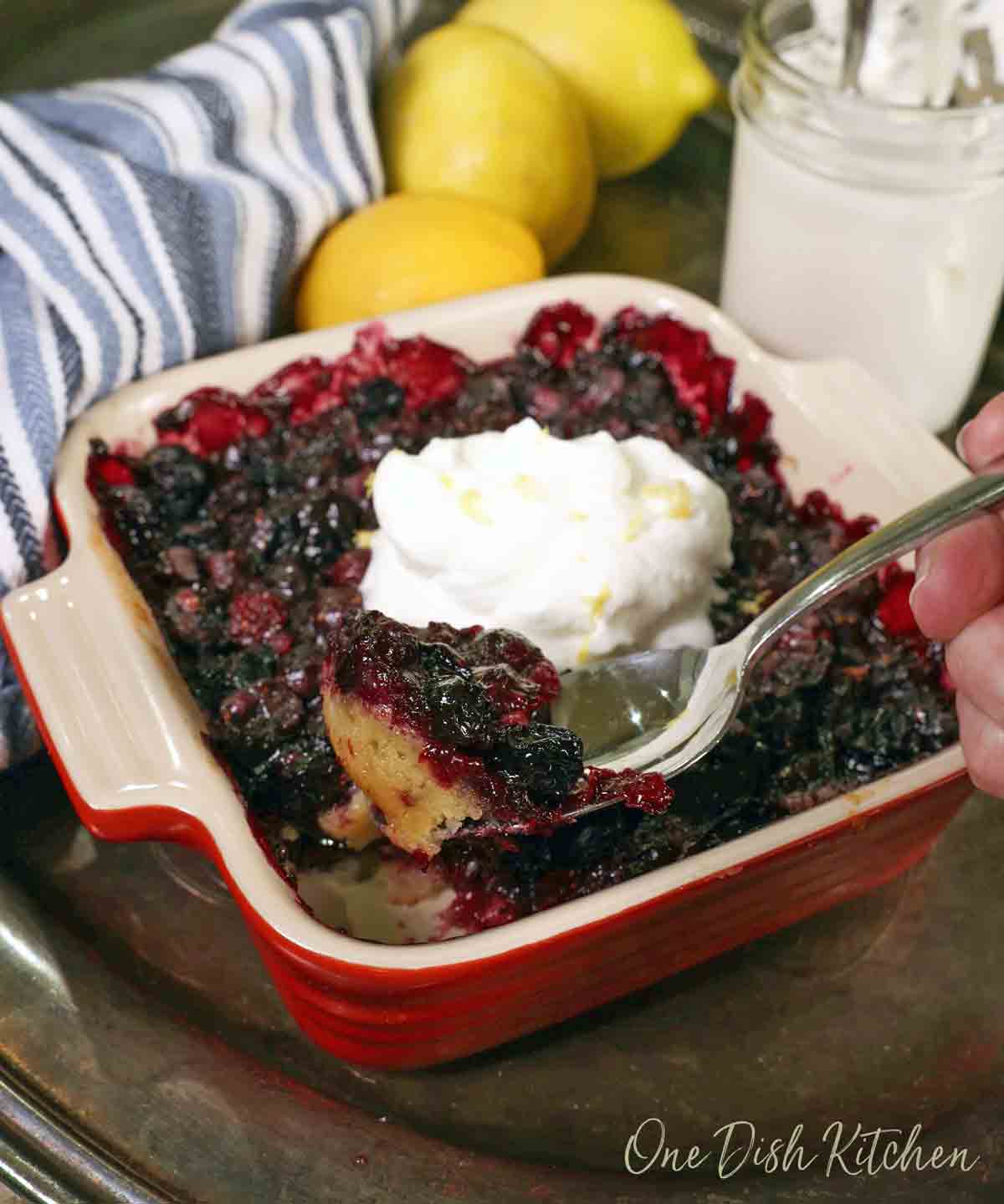 a mini blueberry pie topped with whipped cream in a square red baking dish next to a blue dish towel and 3 lemons.