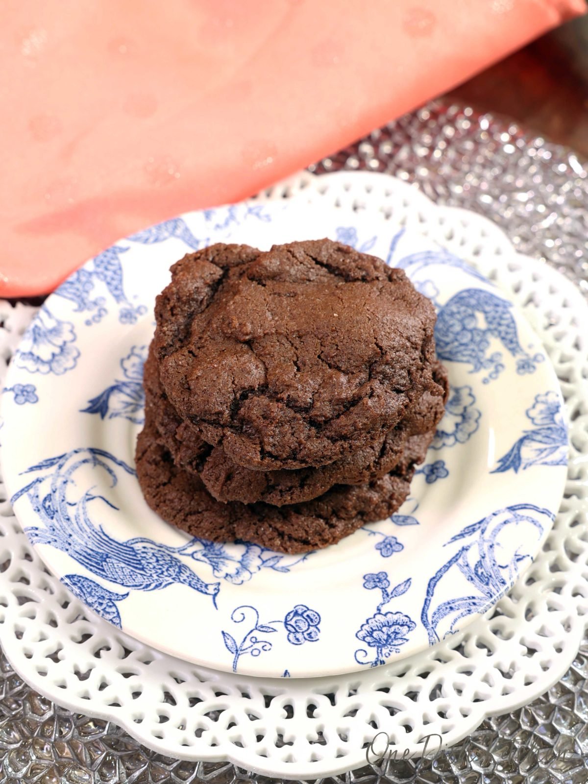 3 chocolate cookies stacked one on top of the other on a blue and white plate next to a peach colored napkin