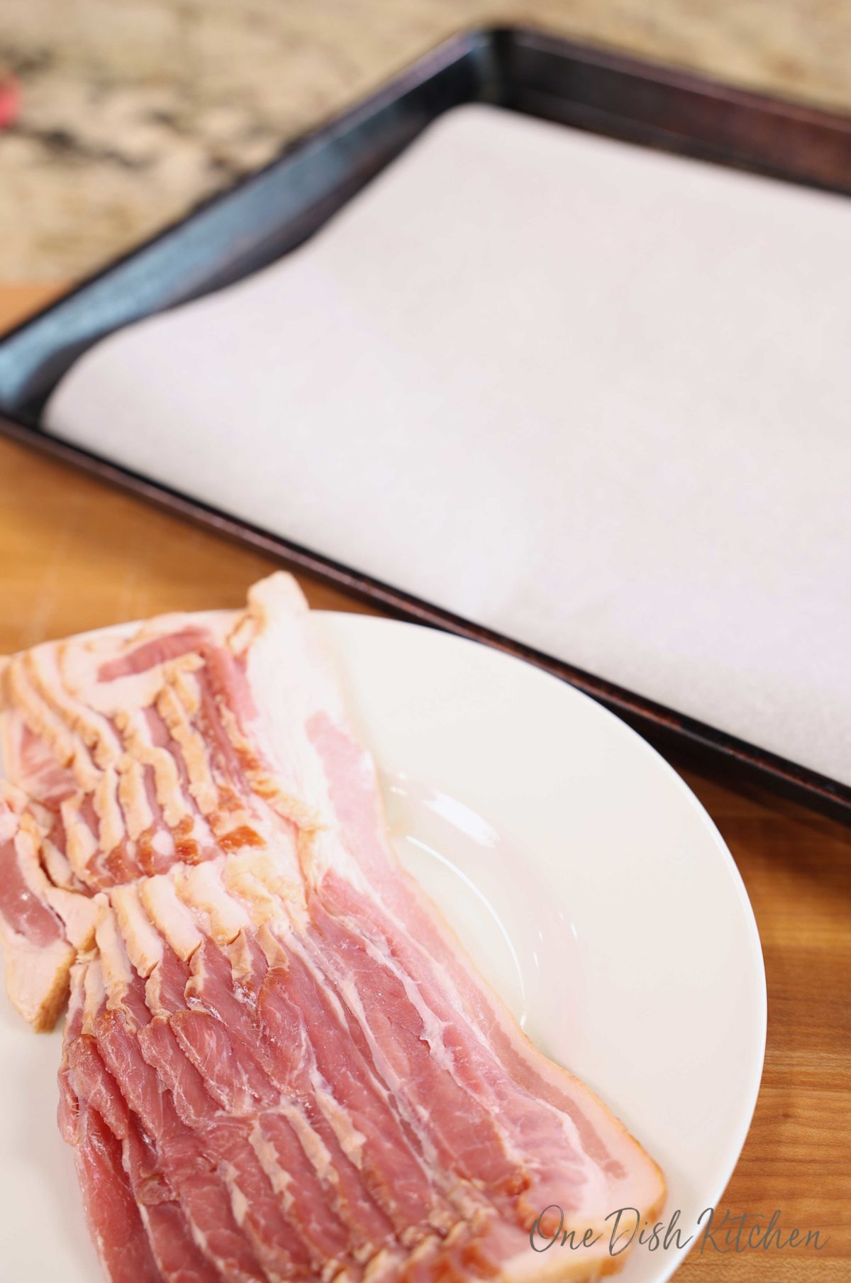 bacon strips on a white plate next to a parchment paper lined baking sheet
