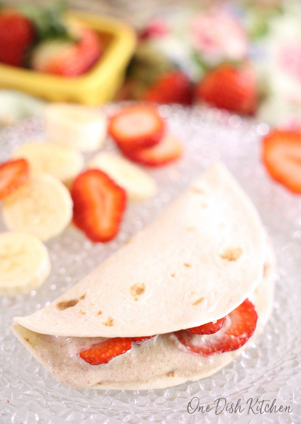 one tortilla folded over and filled with strawberries on a plate with sliced bananas and strawberries surrounding it.