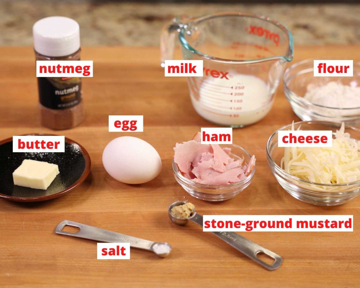 Ingredients needed for a savory dutch baby. milk, egg, ham, cheese, spices, butter and mustard on a brown cutting board.