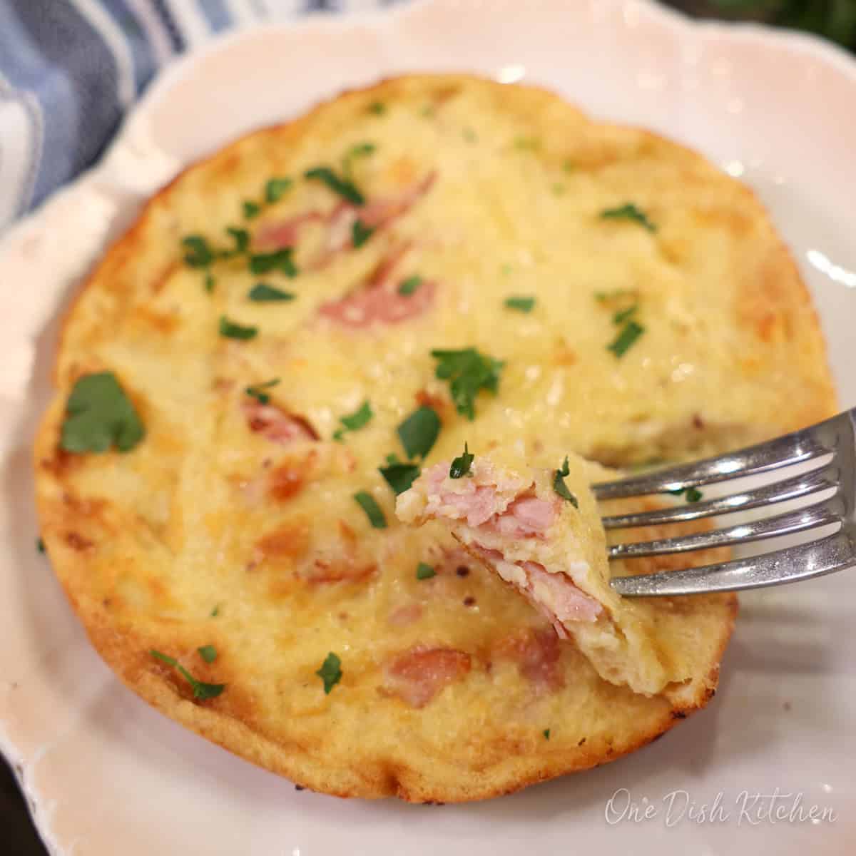 a small ham and cheese dutch baby on a white plate with a fork on the side.