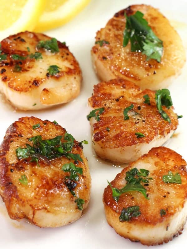 5 pan seared scallops on a white plate next to lemon slices.