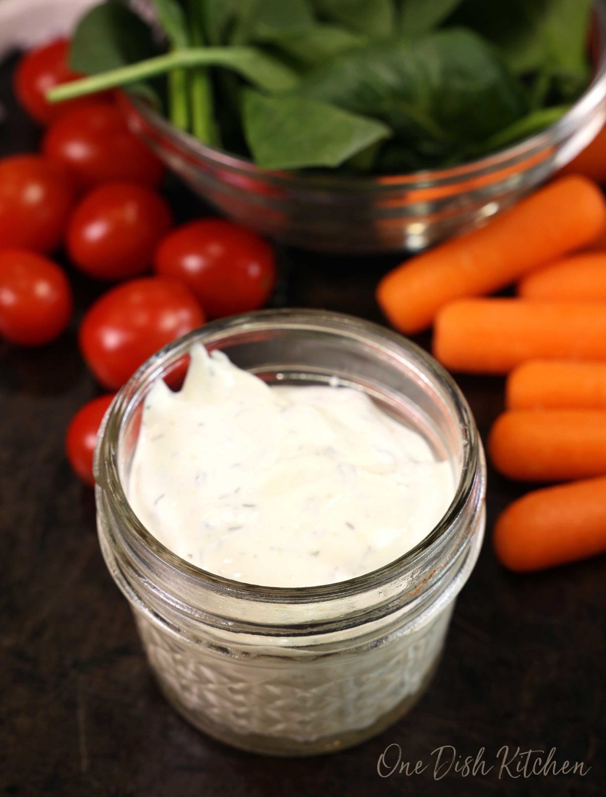 a small clear glass jar of ranch salad dressing next to cherry tomatoes, carrot sticks, and a bowl of spinach.