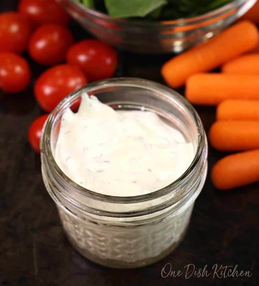 a small clear glass jar of ranch salad dressing next to cherry tomatoes, carrot sticks, and a bowl of spinach.