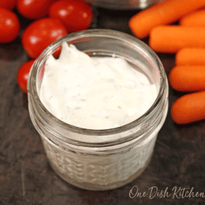 a small mason jar filled with ranch dressing on a black table next to carrots and grape tomatoes.