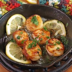 five pan seared scallops in a mini cast iron skillet next to sliced lemons and topped with chopped parsley.