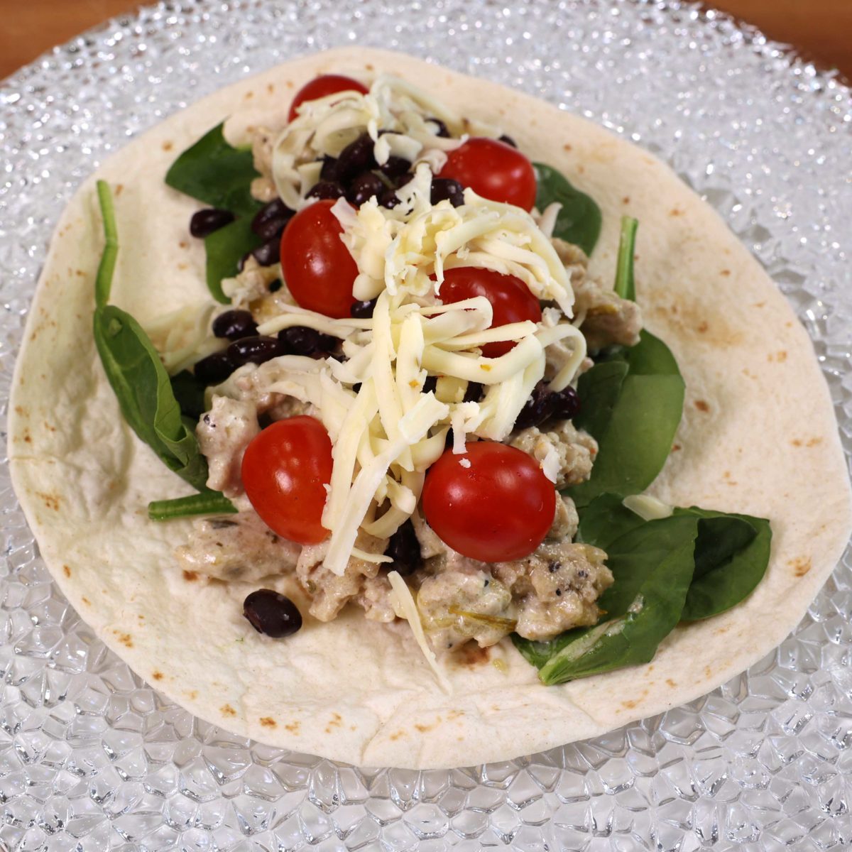 a flour tortilla flat on a plate topped with spinach, ground chicken tomatoes, black beans, and shredded cheese.