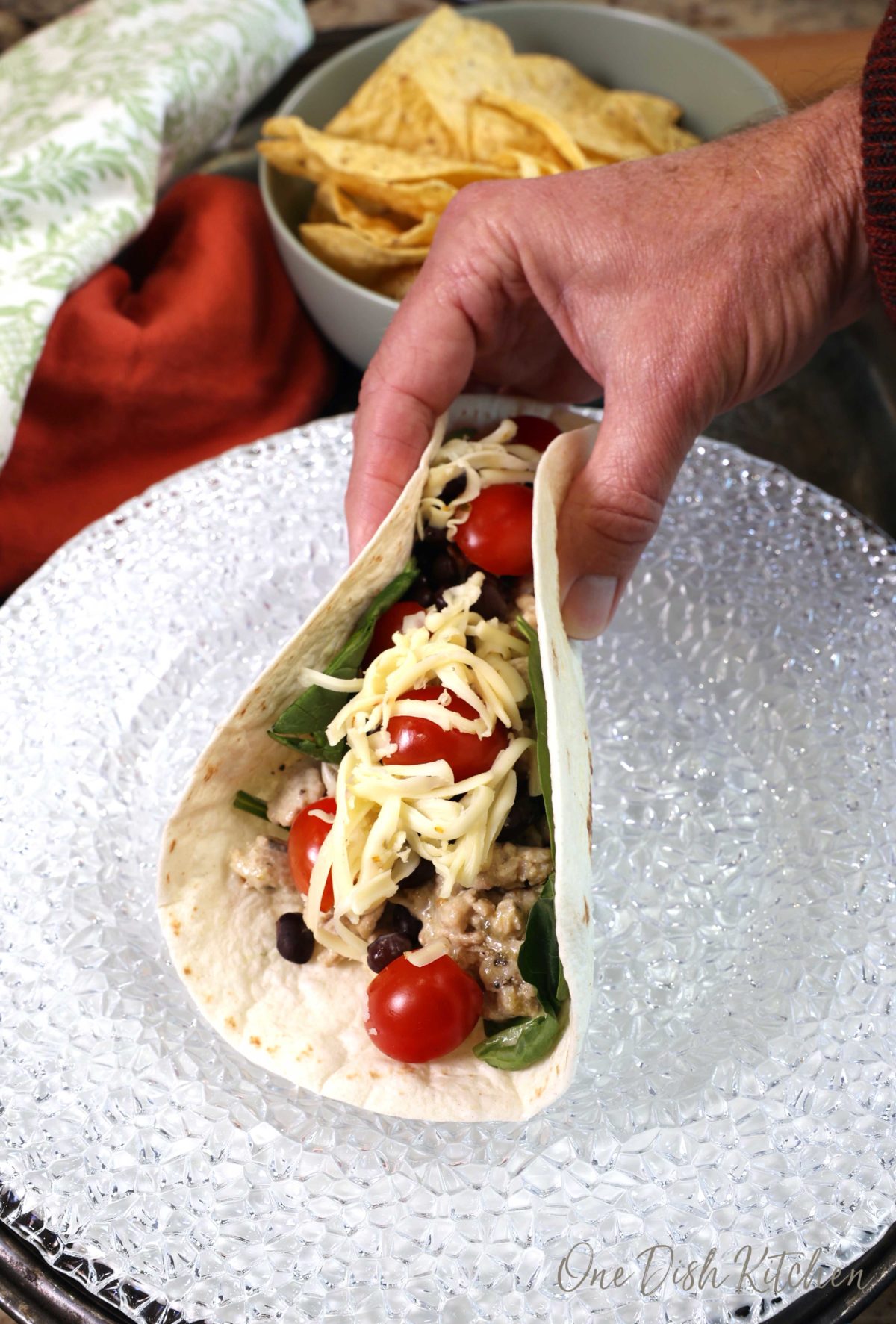 a hand holding a ground turkey wrap topped with spinach, cheese, and tomatoes over a white plate.