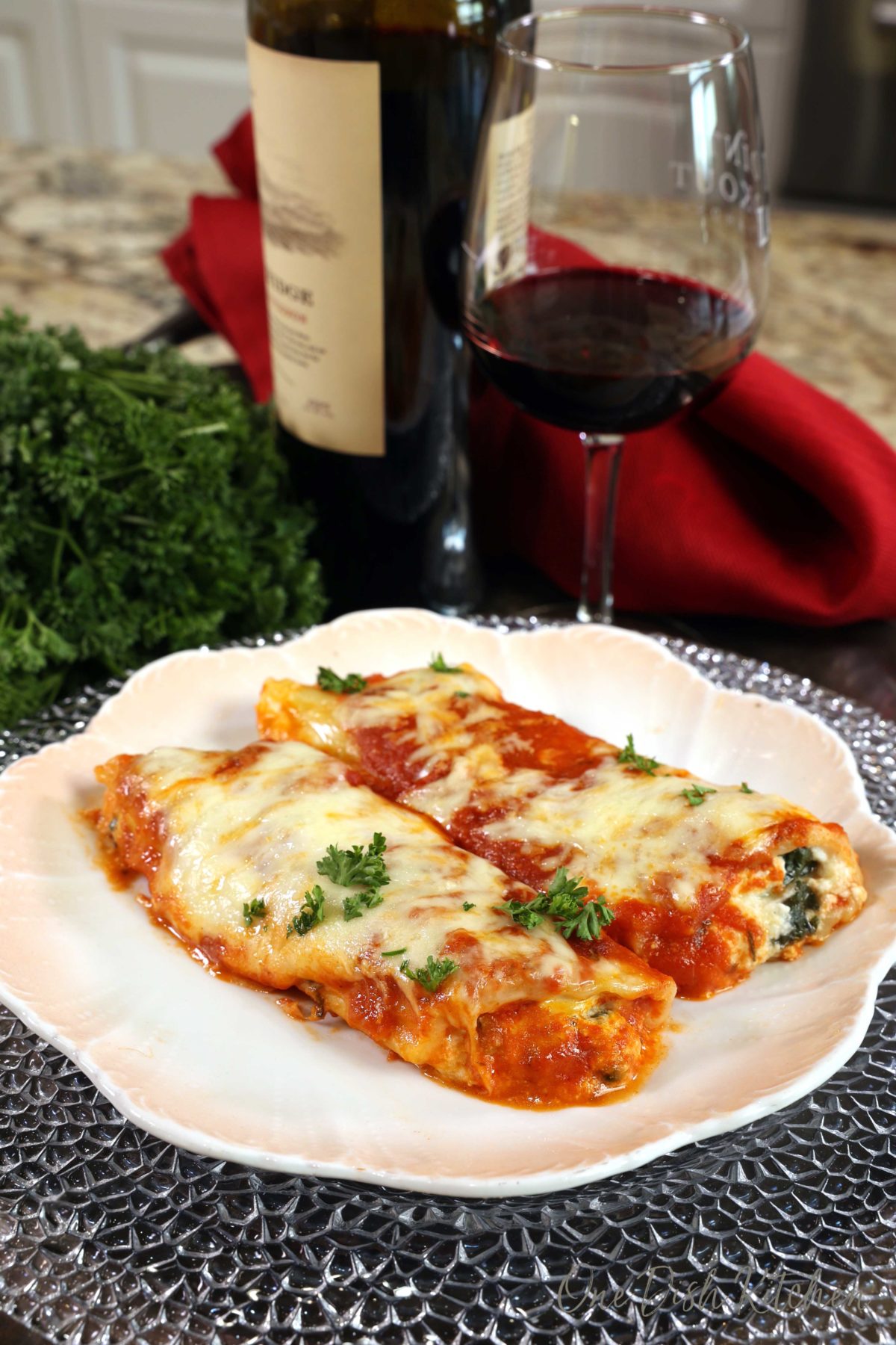 two stuffed manicotti shells on a white plate next to a bottle of red wine.