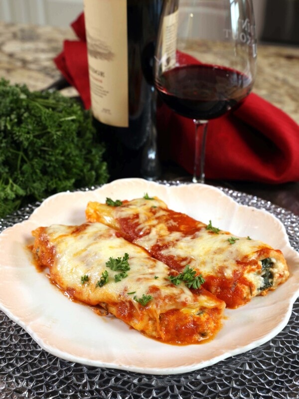 two stuffed manicotti shells on a white plate next to a bottle of red wine