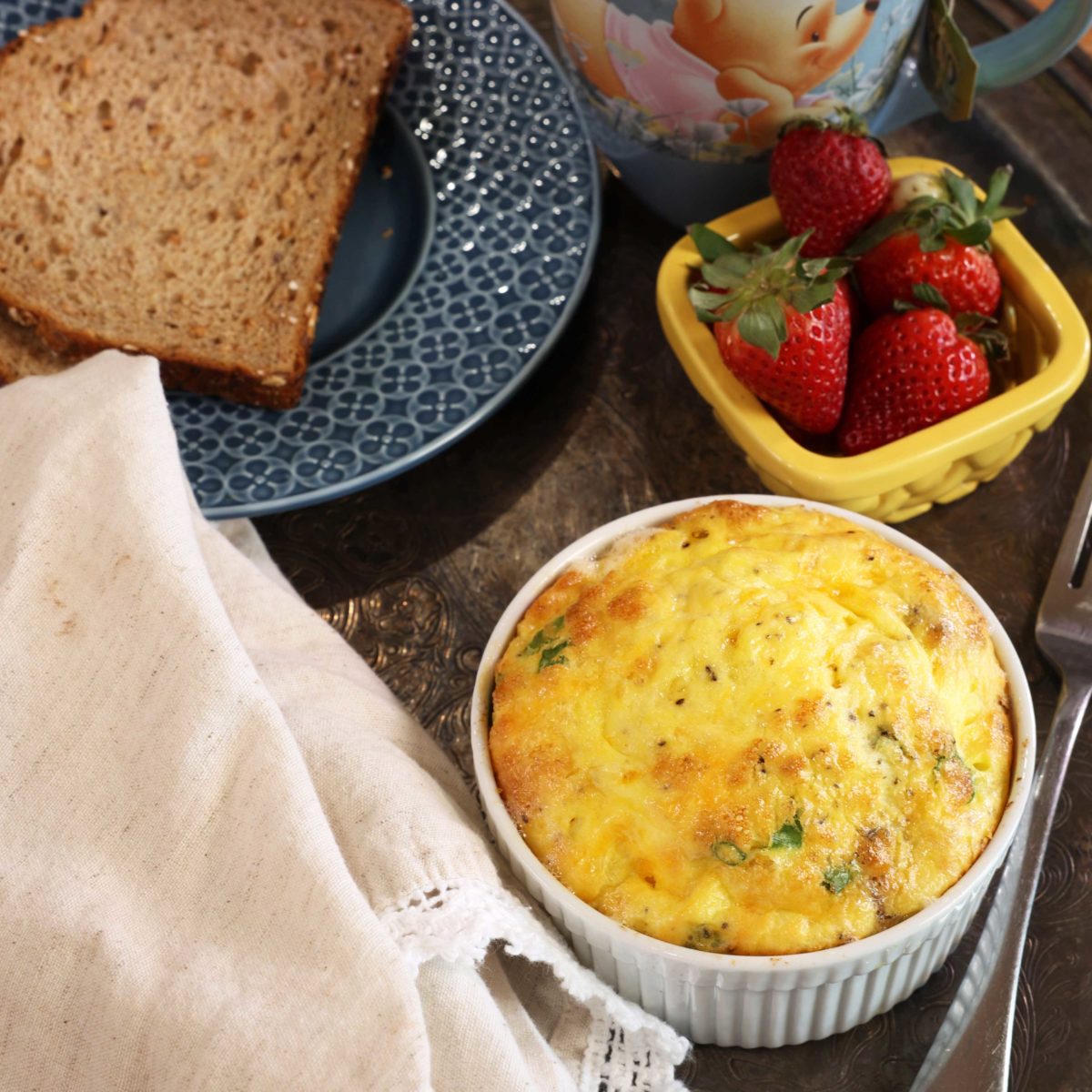 an overhead photo of baked eggs and cheese in a cup next to a blue plate with a slice of toast and a yellow bowl of strawberries.