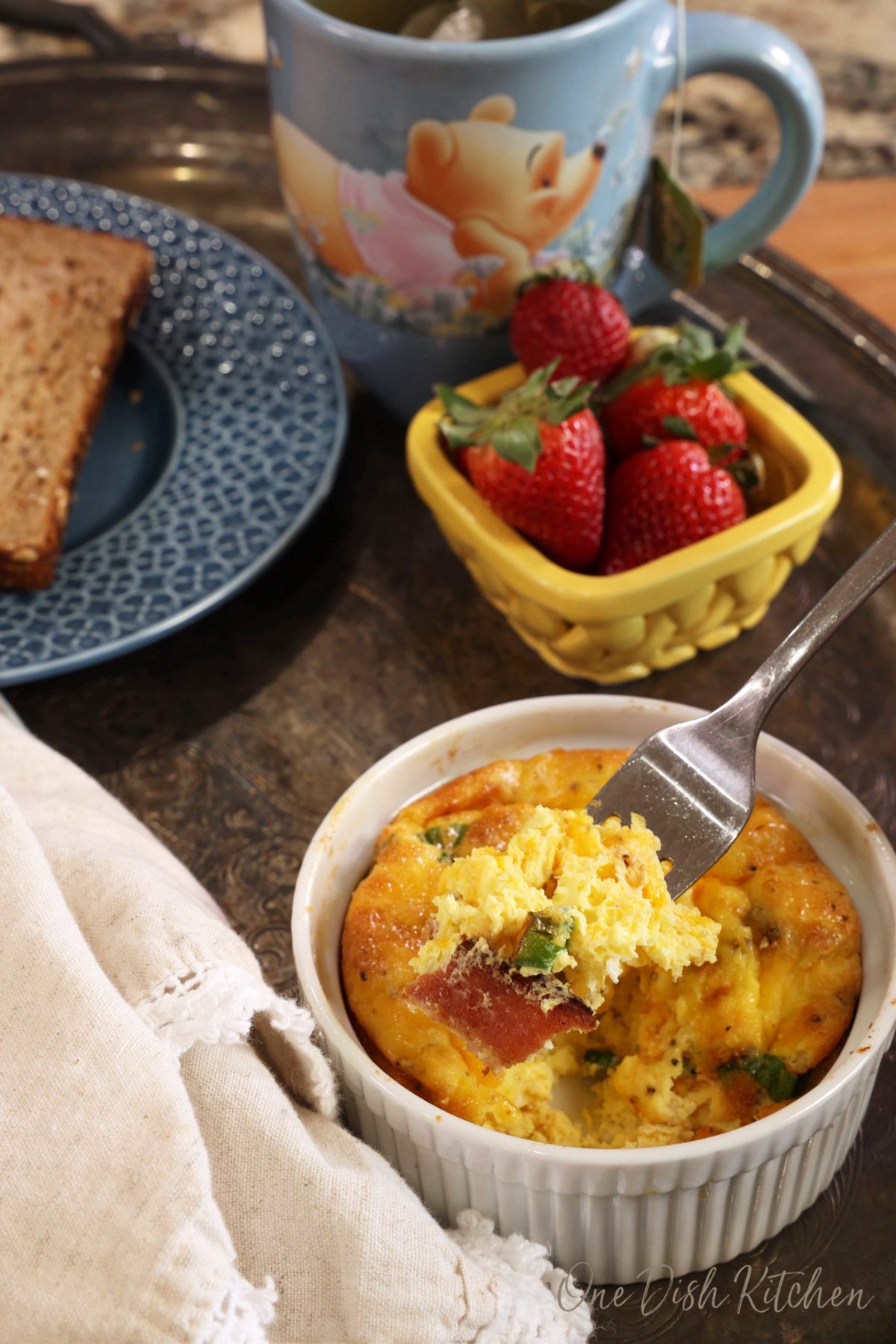 a fork filled with eggs and bacon above a ramekin with baked eggs inside it and next to a bowl of strawberries and a cup of tea.