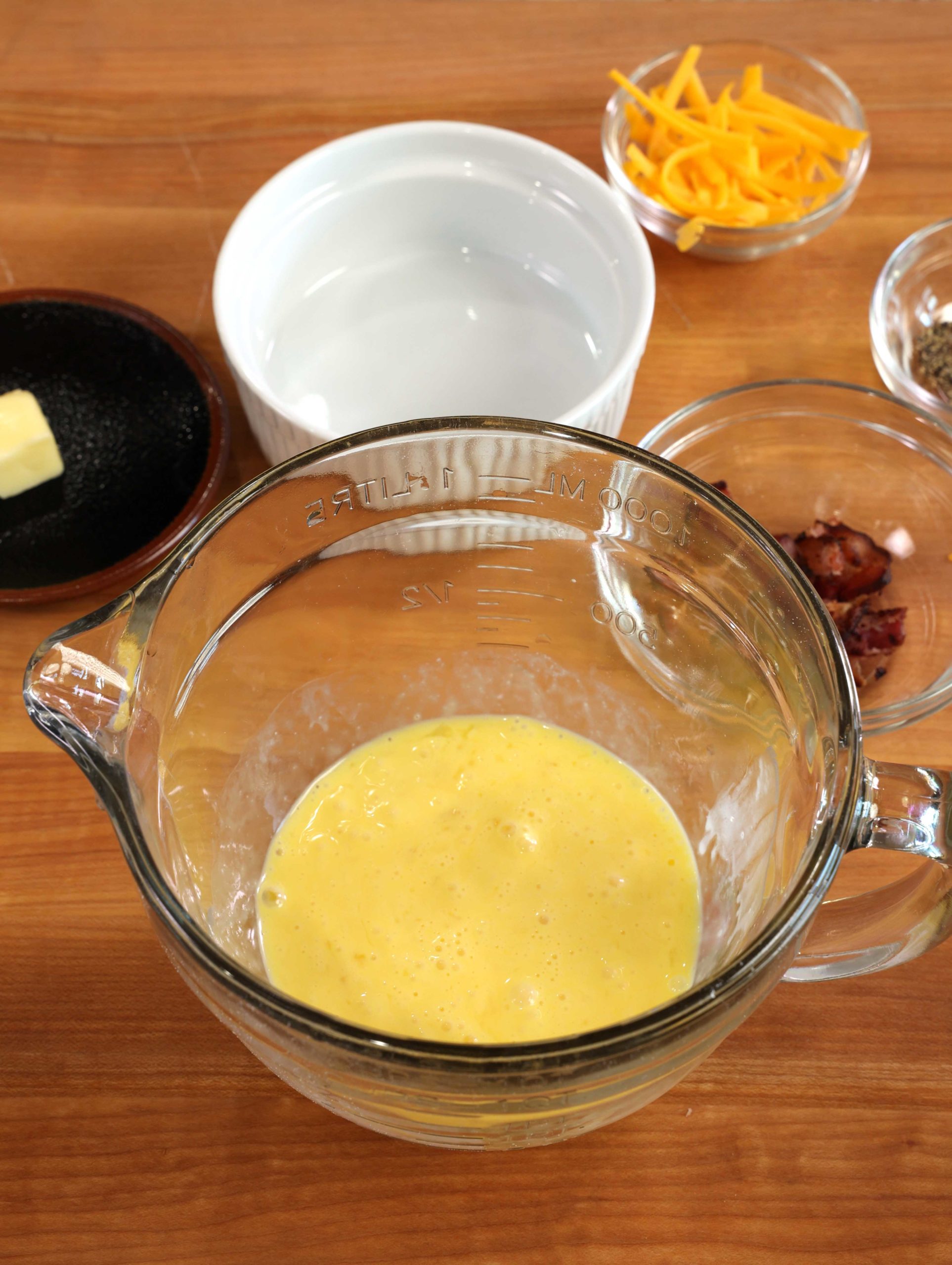 scrambled eggs in a mixing bowl next to bowls of cooked bacon, cheese, butter and a small white ramekin.