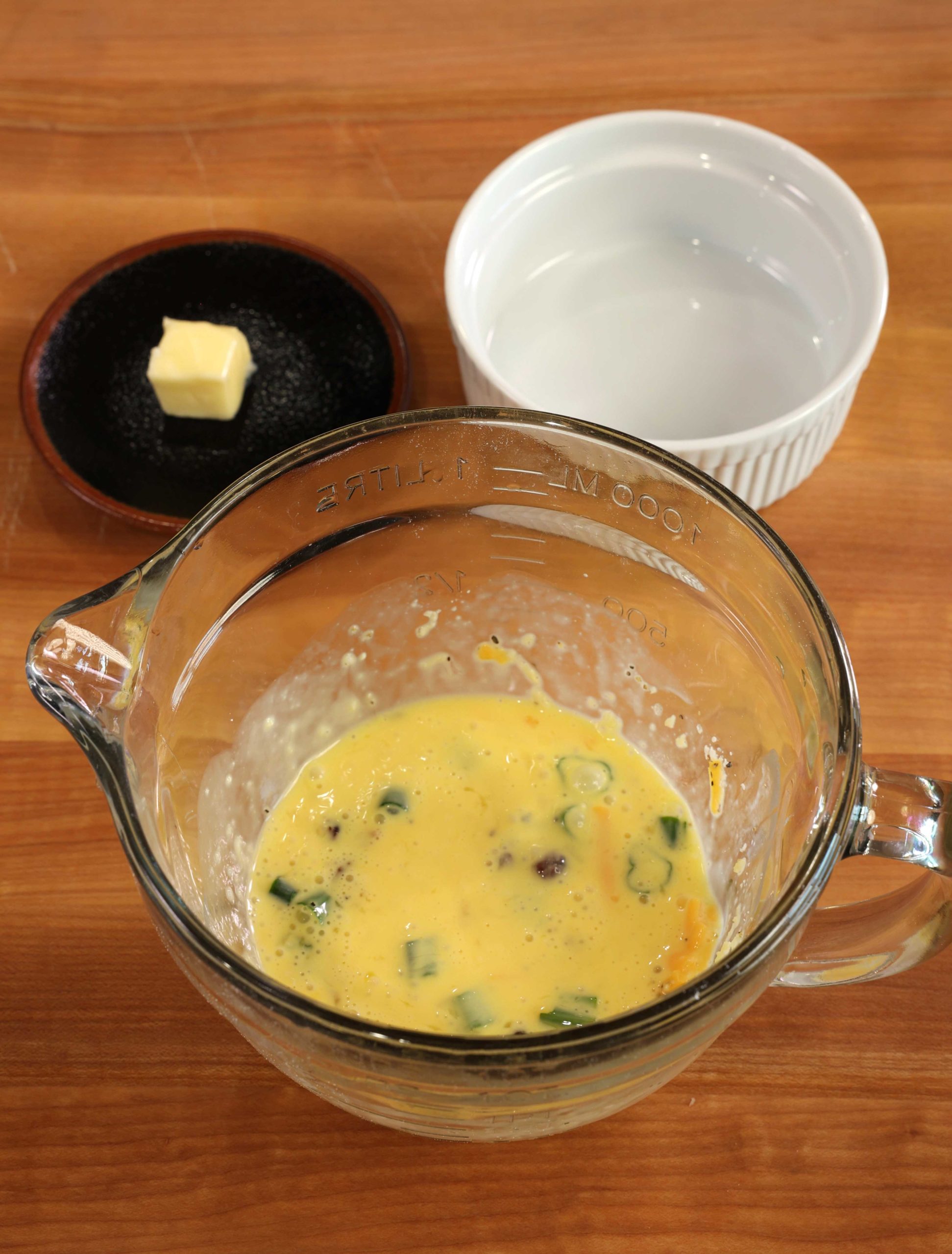 a clear mixing bowl filled with scrambled eggs, bacon, cheese, and green peppers next to a black plate with a small amount of butter on it and a white ramekin.