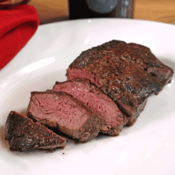 a beef tenderloin sliced on a white plate next to a bottle of red wine and a red napkin.