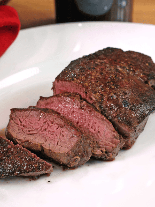 a beef tenderloin sliced on a white plate next to a bottle of red wine and a red napkin.