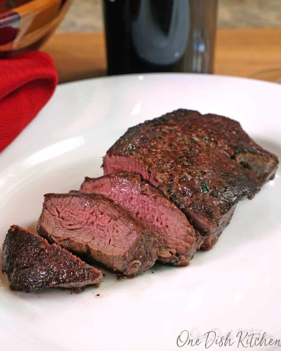 a beef tenderloin sliced on a white plate next to a bottle of red wine and a red napkin.