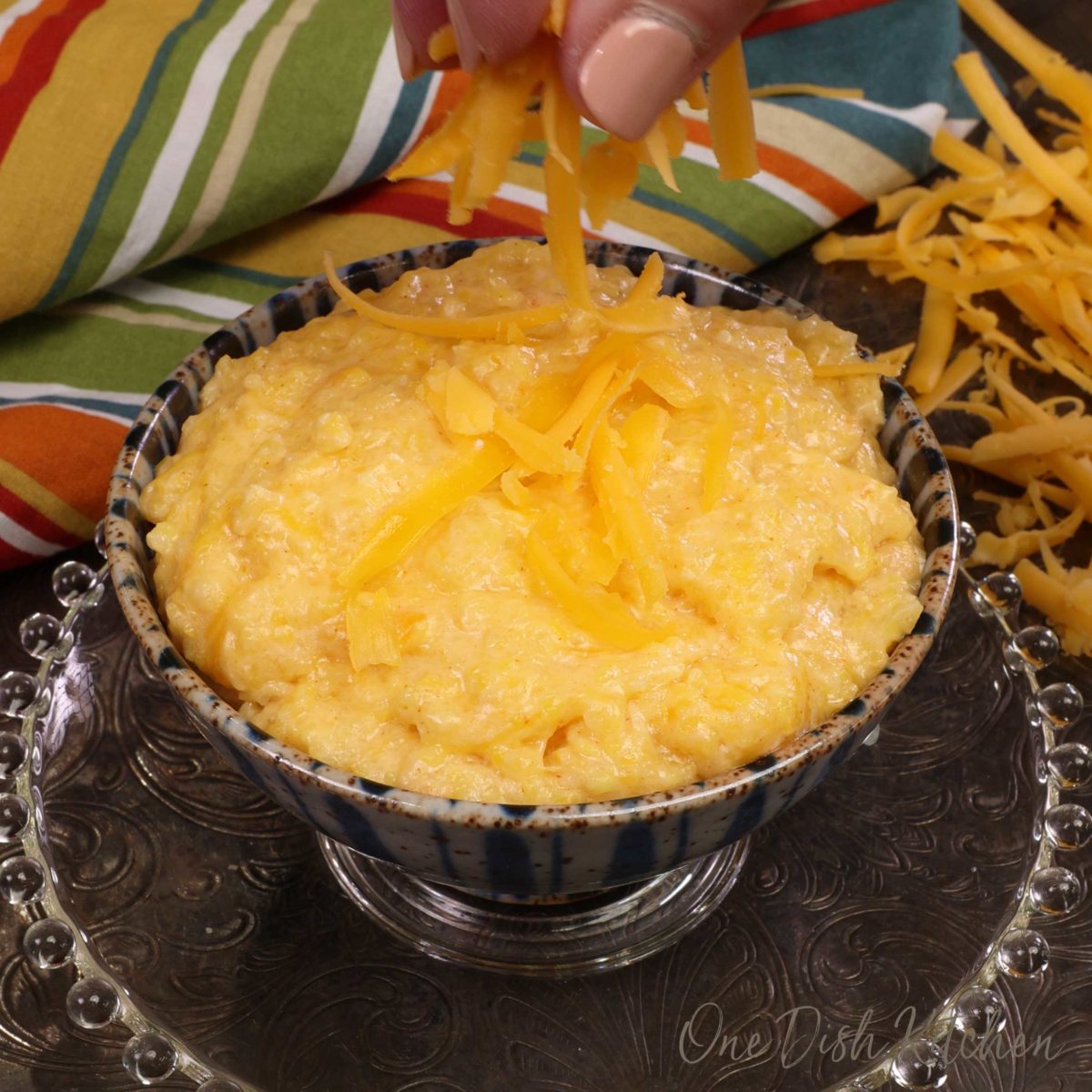 a small blue bowl filled with cheese grits with a hand sprinkling extra cheese over the top.