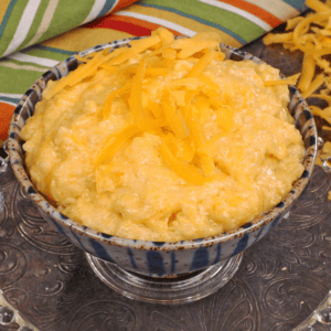 a small bowl of cheese grits next to shredded cheese and a large block of cheese in the background.
