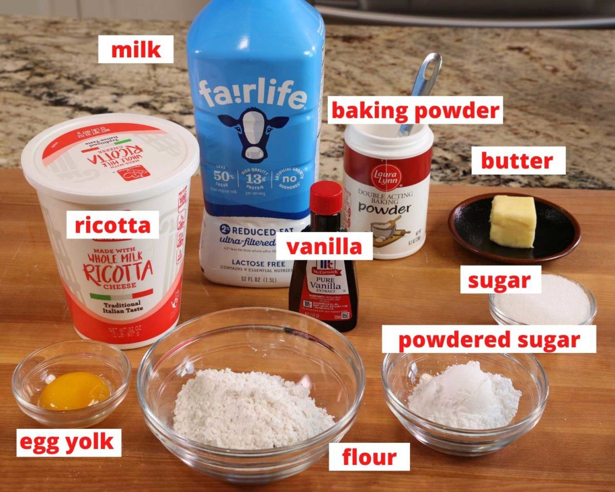 milk, flour, sugar, baking powder, ricotta cheese, an egg and vanilla on a wooden cutting board.